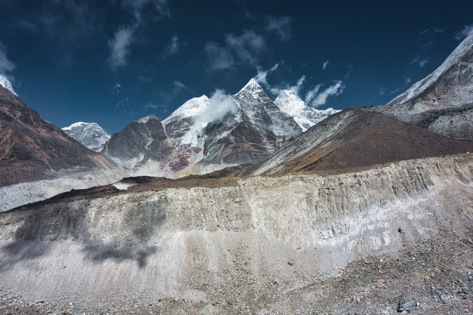 Gebirgslandschaft mit schneebedeckten Gipfeln und kargen Hängen unter blauem Himmel.