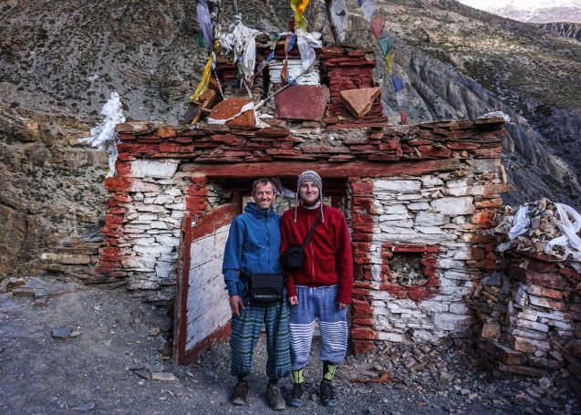 Zwei Personen vor einem kleinen, baufälligen Gebäude in gebirgiger Umgebung.