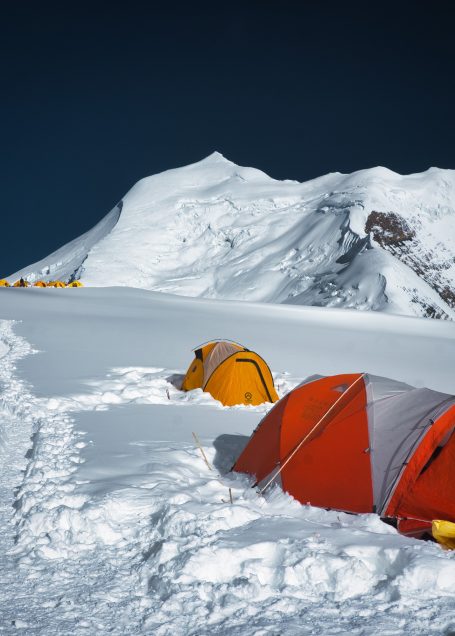 Zwei orangefarbene Zelte auf schneebedecktem Boden vor einem Berggipfel.