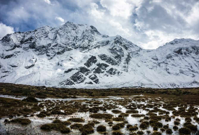 Schneebedeckte Berge unter einem bewölkten Himmel, mit grünem Gras im Vordergrund.