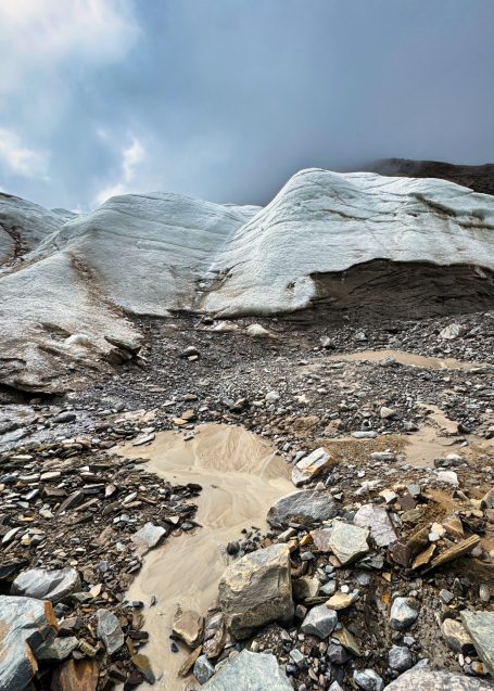 Gletscher mit Stein- und Schuttansammlungen im Vordergrund, grauer Himmel im Hintergrund.