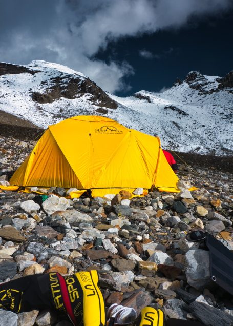 Gelbes Zelt auf steinigem Boden, im Hintergrund schneebedeckte Berge und dunkler Himmel.