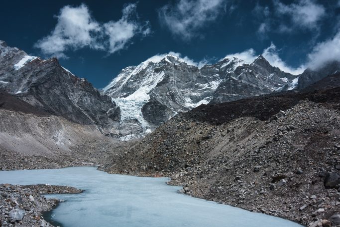 Berglandschaft mit Gletscher und blauem See im Vordergrund, unter einem bewölkten Himmel.