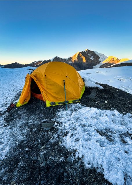 Gelbes Zelt auf schneebedecktem Boden mit Bergen im Hintergrund bei Sonnenaufgang.