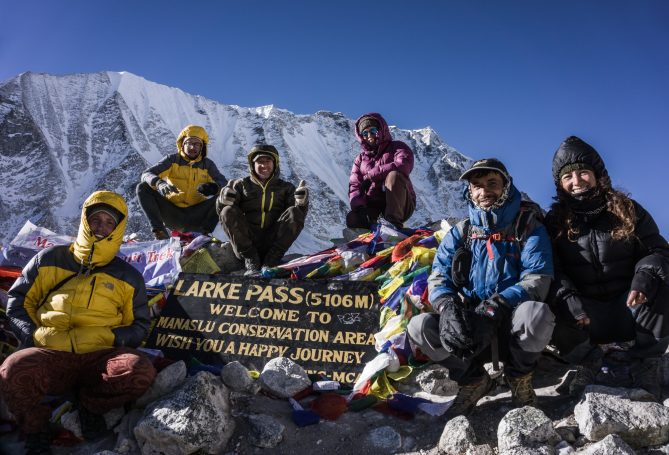 Sechs Personen stehen und sitzen auf einem Berggipfel mit Fahnen und einem Begrüßungsschild.