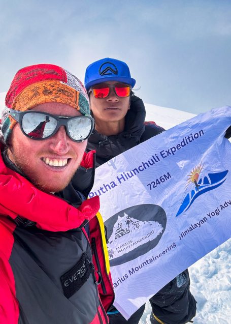Zwei Personen posieren auf einem Berggipfel mit einem Flaggenbanner.