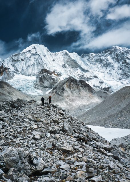 Zwei Wanderer auf einem steinigen Pfad vor schneebedeckten Bergen und bewölktem Himmel.