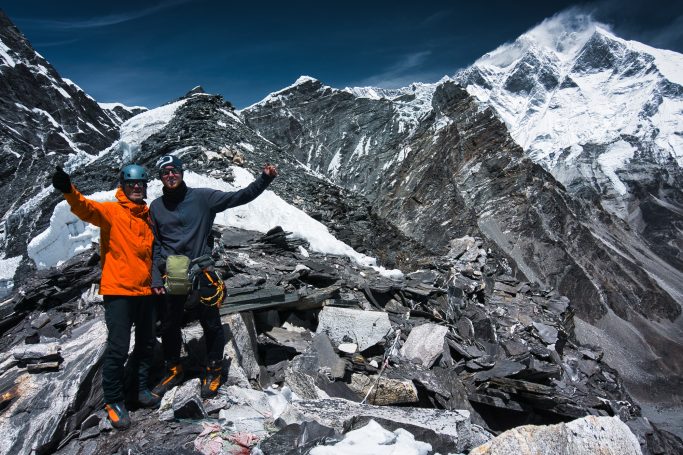 Zwei Bergsteiger posieren jubelnd auf einem schneebedeckten Gipfel mit Bergen im Hintergrund.