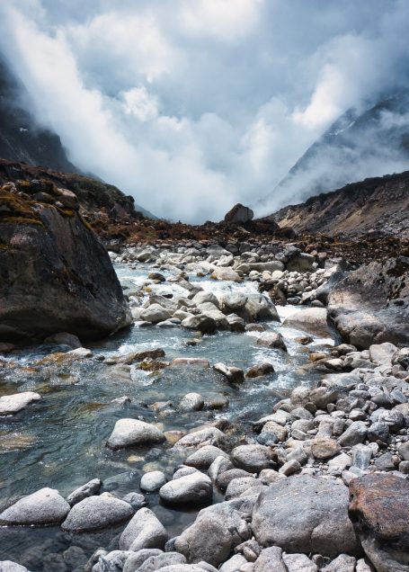 Berglandschaft mit einem klaren Fluss, umgeben von Felsen und dichten Wolken.