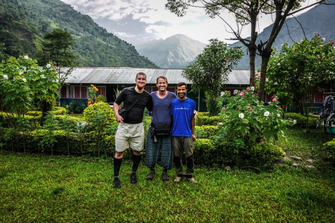 Drei Männer stehen in einem Garten mit Bergen im Hintergrund. Grüne Pflanzen und Blumen sind zu sehen.