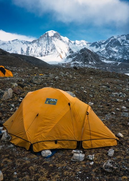 Gelbes Zelt in gebirgiger Landschaft mit schneebedeckten Gipfeln im Hintergrund.