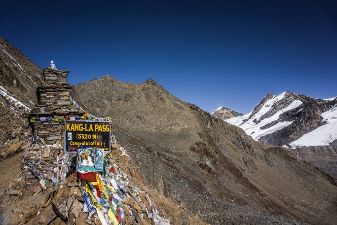 Gebirgspass mit bunten Gebetsfahnen und einem Schild, umgeben von schneebedeckten Gipfeln.