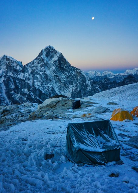Schneebedeckte Berge mit Zelten vor einem klaren, blauen Himmel und einem Mond.