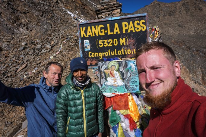 Drei Personen stehen am Kang-La Pass-Schild, umgeben von Felsen und bunten Tüchern.