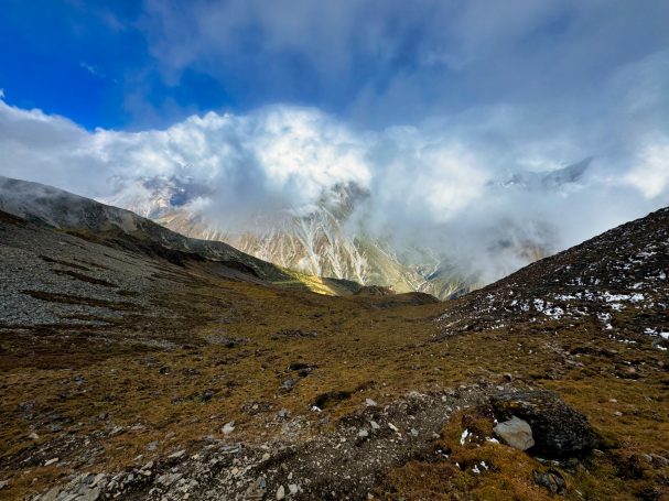 Berglandschaft mit bewölktem Himmel und grün-braunem Gelände im Vordergrund.