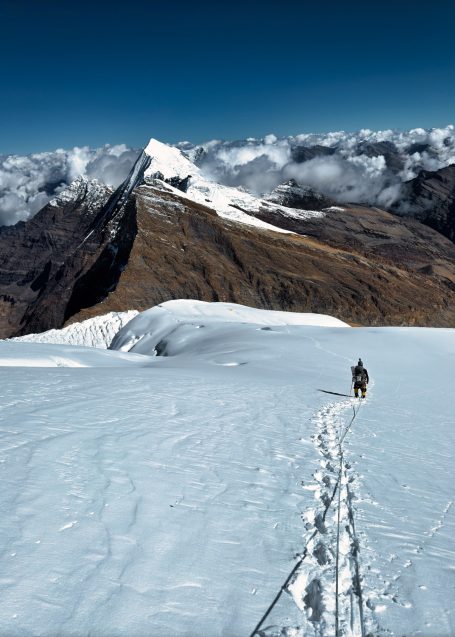Ein Bergsteiger wandert auf schneebedecktem Gelände mit dramatischem Bergpanorama im Hintergrund.