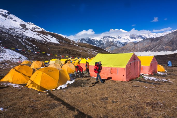 Berghütte und Gelbe Zelte in einer Gebirgslandschaft mit schneebedeckten Gipfeln.