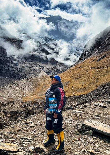 Person in Wanderkleidung steht in einer bergigen Landschaft mit Wolken und Felsen.