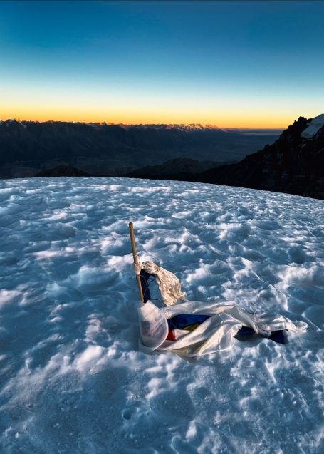 Ein Bergsteiger liegt im Schnee mit einem Bergpanorama und einem Sonnenuntergang im Hintergrund.