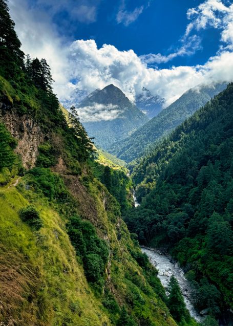 Berglandschaft mit Fluss und Wolken, umgeben von grünen Wäldern und Gipfeln.