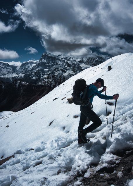 Bergsteiger mit Rucksack, der einen schneebedeckten Hang erklimmt, bewölkter Himmel.