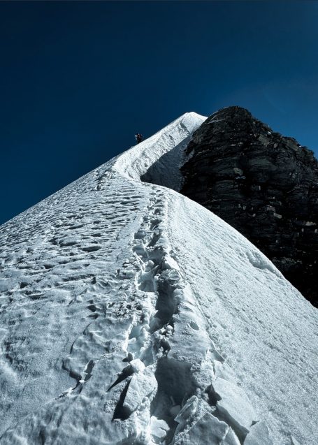 Ein schmaler, verschneiter Grat führt zu einem hohen Gipfel unter blauem Himmel.