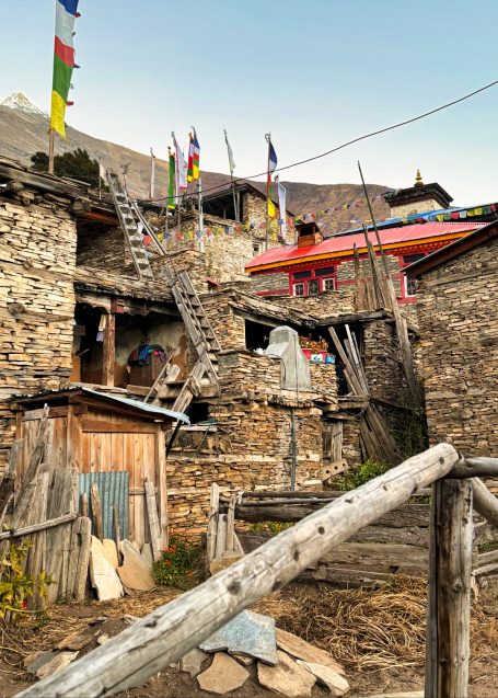 Traditionelles Dorf mit Steinhäusern, Holzleitern und bunten Fahnen im Hintergrund.