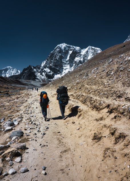 Zwei Wanderer mit Rucksäcken gehen einen staubigen Weg in bergiger Landschaft.