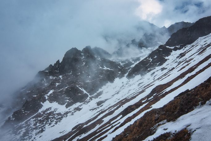 Es zeigt schneebedeckte Berge in nebliger, stürmischer Umgebung.