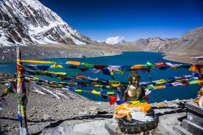 Buddha-Statue mit bunten Gebetsfahnen am See, umgeben von schneebedeckten Bergen.