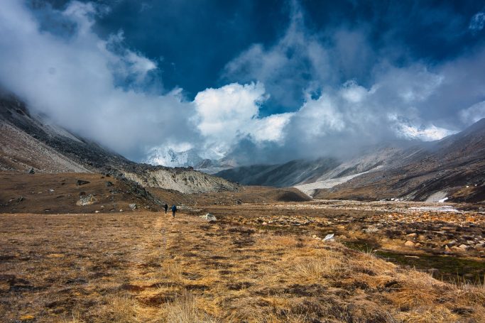 Berglandschaft mit Wolken und kargem Gelände, Wanderer im Hintergrund.