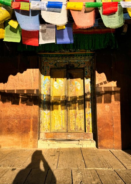 Bunte Gebetsfahnen hängen über einer alten, gelben Tür aus Holz.